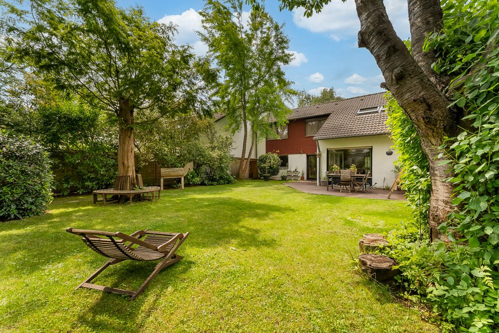 Grüner Garten mit Bäumen, Liege, Bank und Terrasse an einem Wohnhaus.
