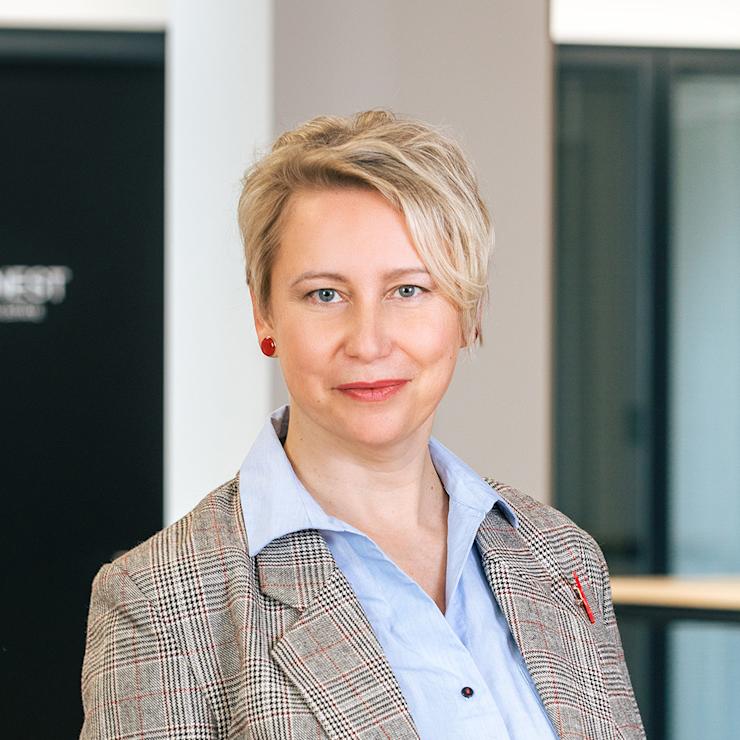 Person mit kurzem, blondem Haar steht in einem Innenraum mit Glaswänden und Holzboden.