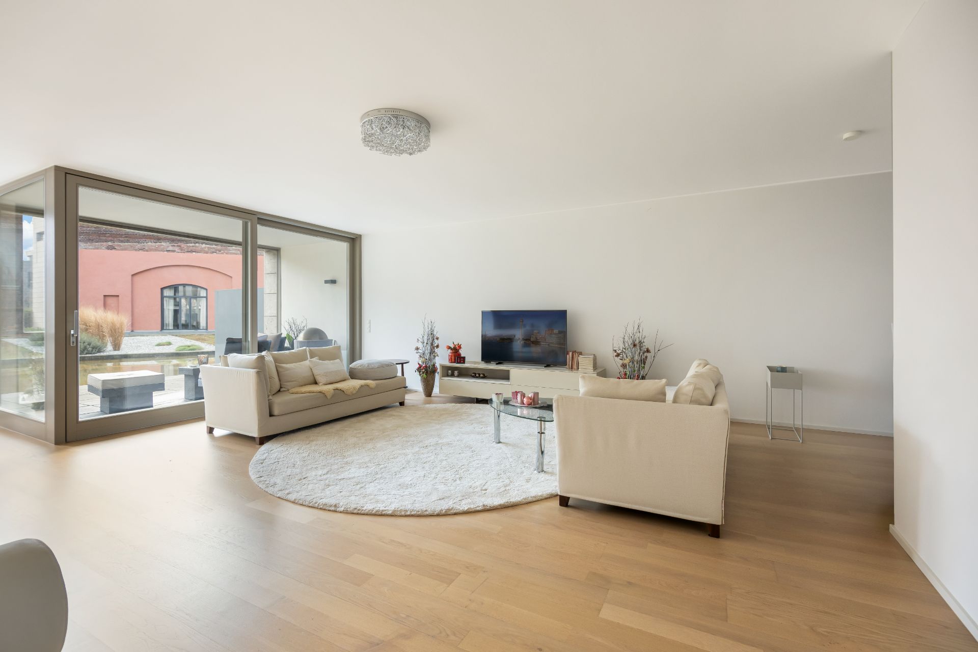 Helles Wohnzimmer mit zwei Sofas, Glastisch, Fernseher und großen Fenstern zum Garten.