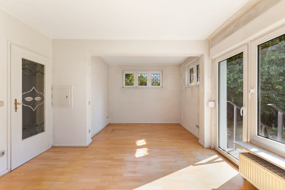 Heller Raum mit Parkettboden, weißen Wänden und großen Fenstern, Blick ins Grüne.