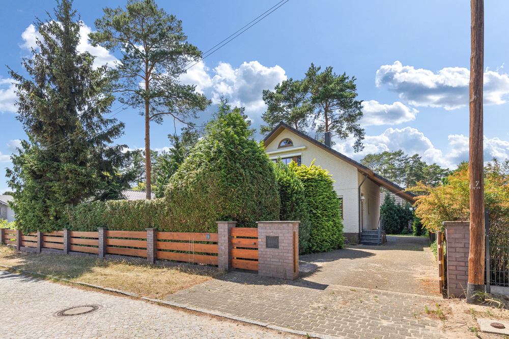 Einfamilienhaus mit Garten, Einfahrt und Holzzaun, von Bäumen und Sträuchern umgeben.