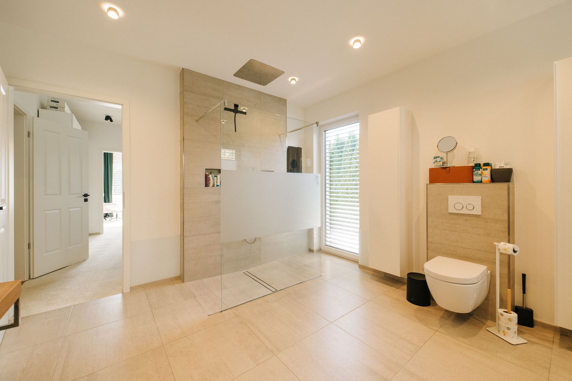 Modernes Badezimmer mit offener Dusche, Hängetoilette und großem Fenster mit Jalousien.
