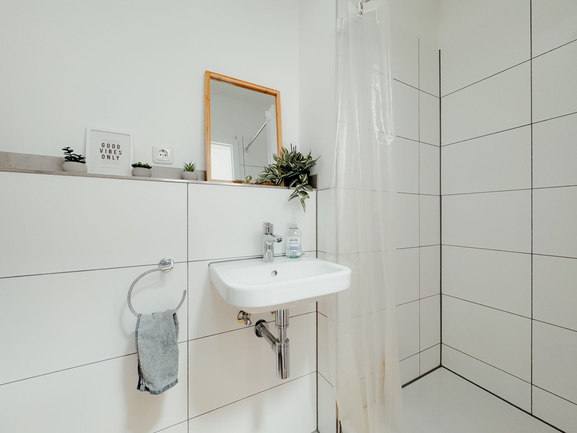 Helles Badezimmer mit Waschbecken, Spiegel, Pflanzen und einer offenen Dusche mit Vorhang.