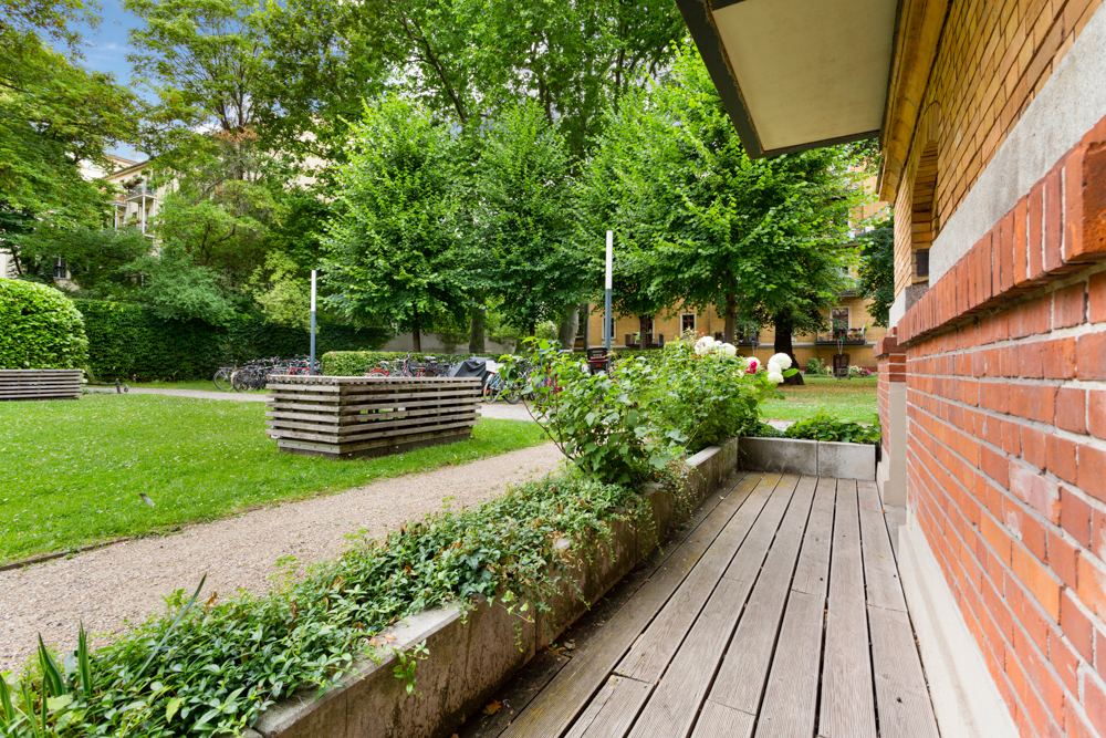Holzterrasse an einem Backsteingebäude mit Blick auf grünen Garten und Fahrradständer.