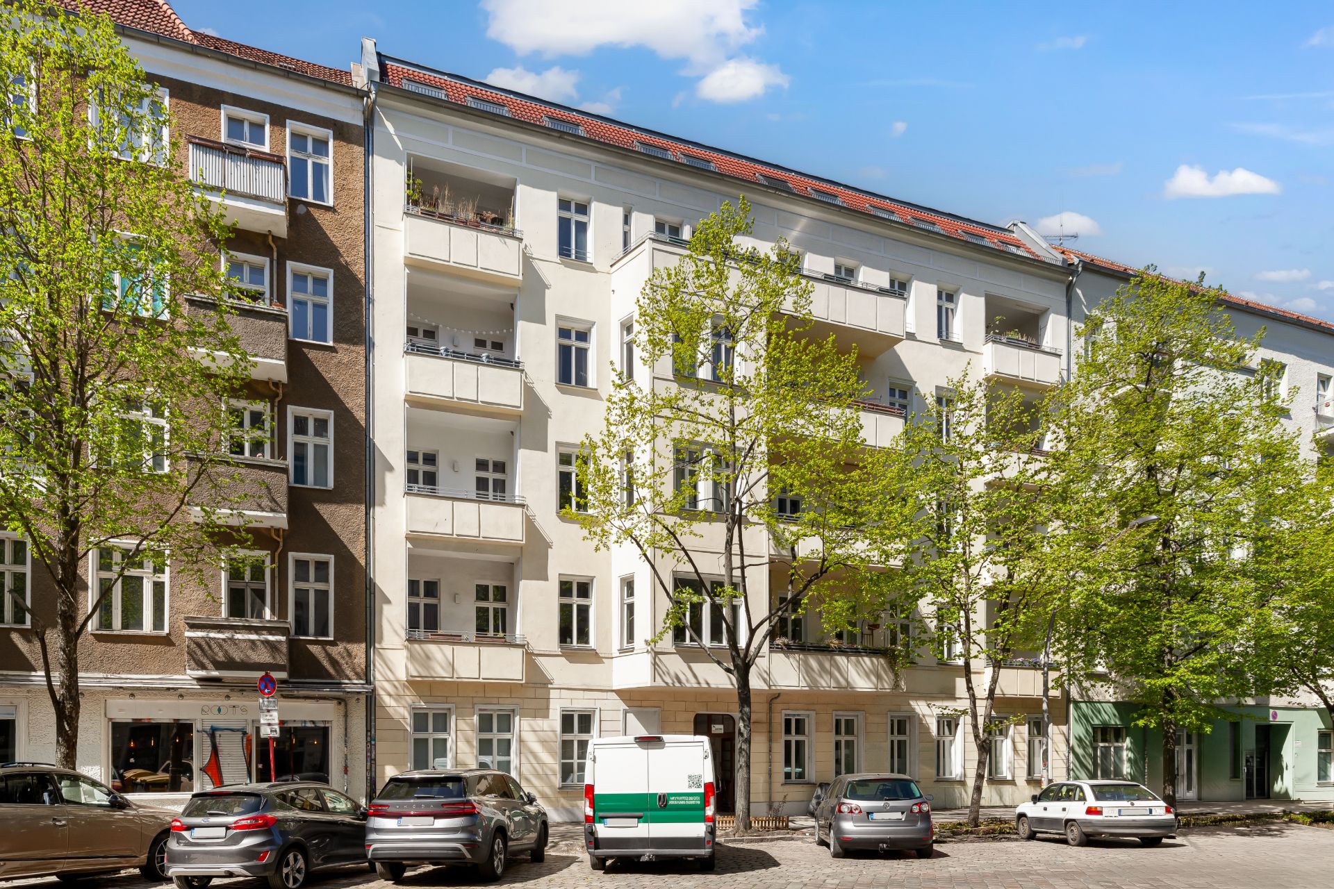 Mehrstöckiges Wohnhaus mit Balkonen und geparkten Autos davor, Bäume an der Straße.