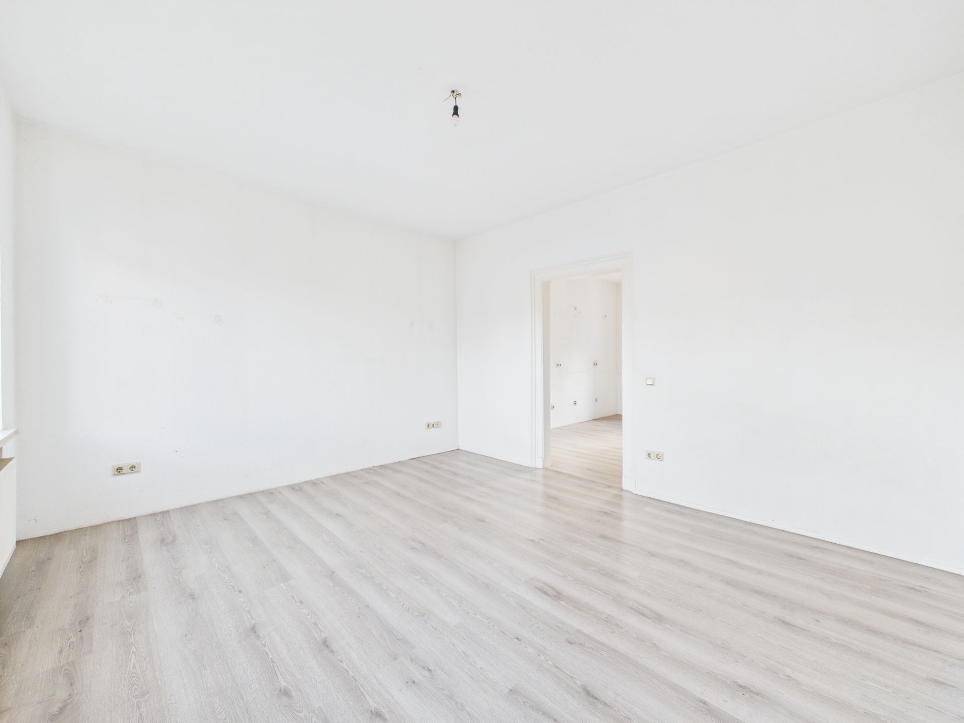 Leerer, heller Raum mit weiß gestrichenen Wänden und hellgrauem Laminatboden.