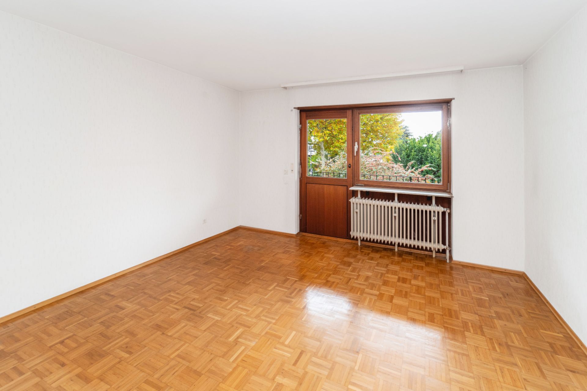 Leerer Raum mit Parkettboden, weißer Wand, Fenster und Heizung unter dem Fenster.