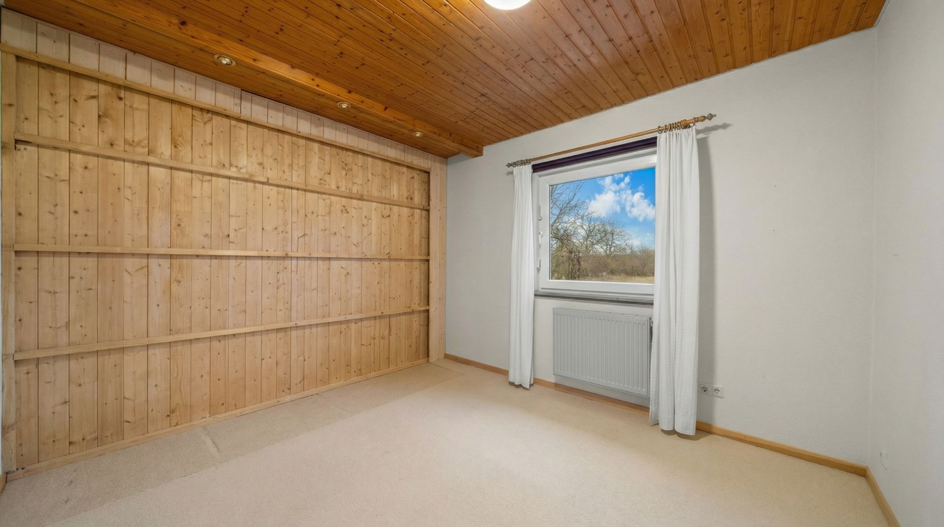Leeres Zimmer mit holzvertäfelter Wand, Holzdecke, Fenster mit weißen Vorhängen und Heizkörper.