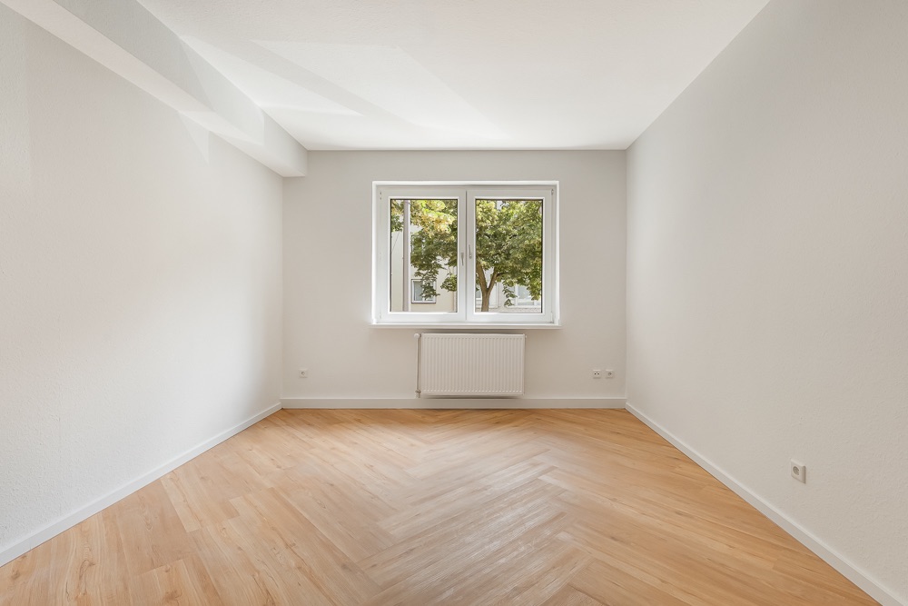 Leerer, heller Raum mit Holzfußboden, weißem Fenster und Heizkörper unter dem Fenster.
