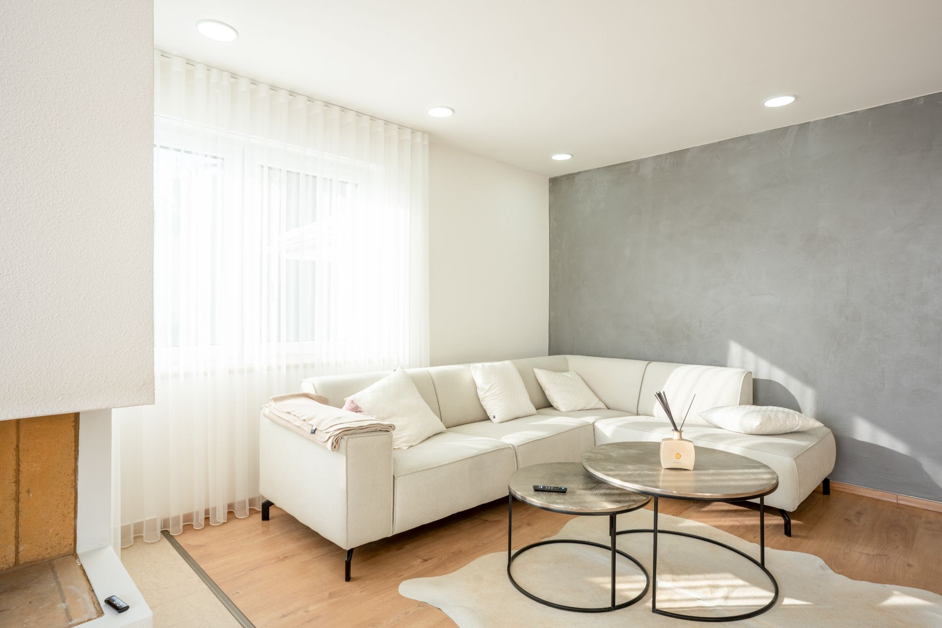 Helles Wohnzimmer mit weißem Ecksofa, Couchtischen und großen Fenstern mit Vorhängen.