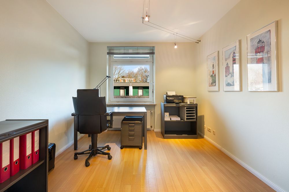 Helles Arbeitszimmer mit Schreibtisch, Bürostuhl, Regalen und Bildern an der Wand.
