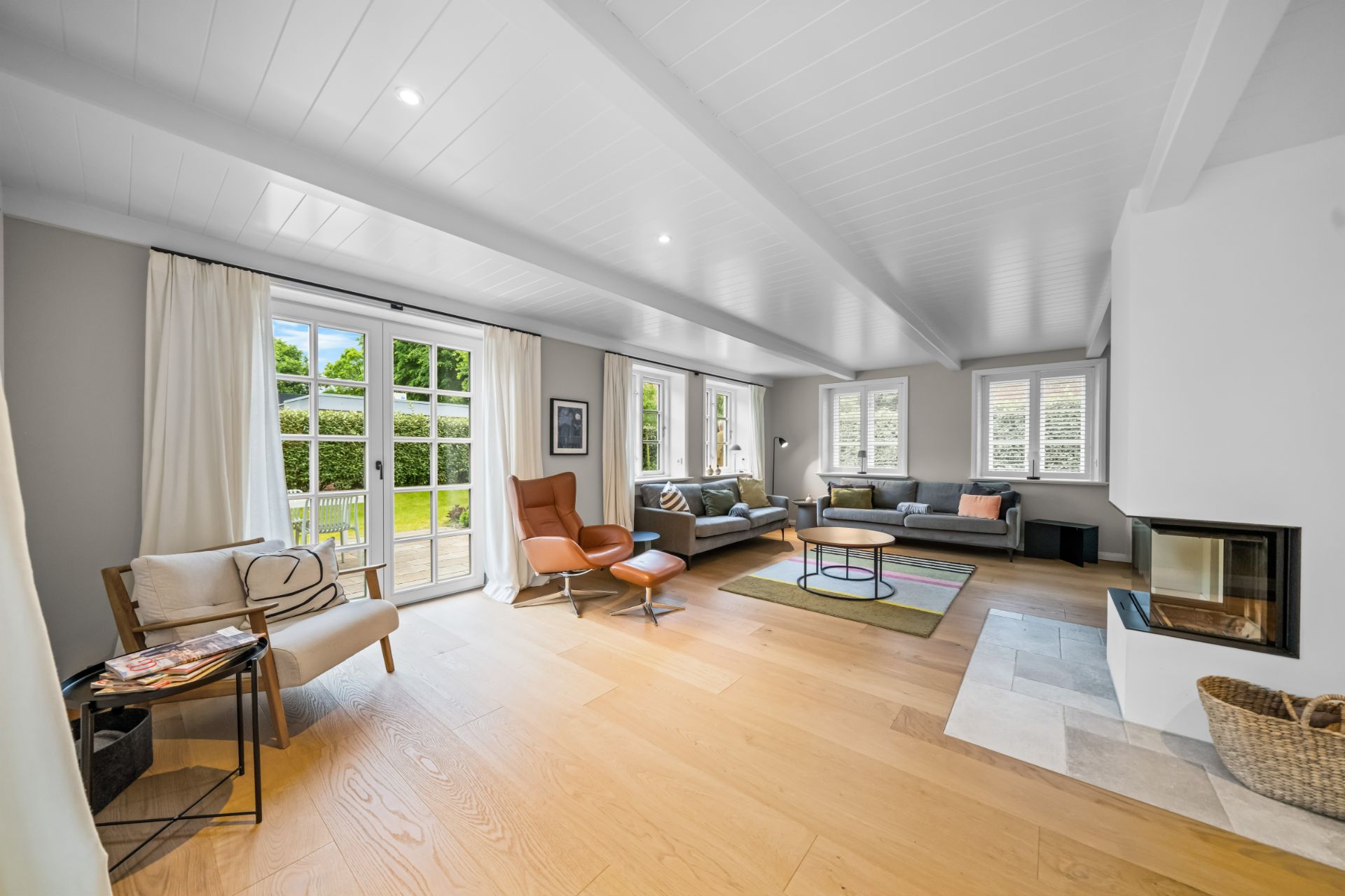 Helles Wohnzimmer mit Sofa, Sesseln, Kamin und großen Fenstern zum Garten.