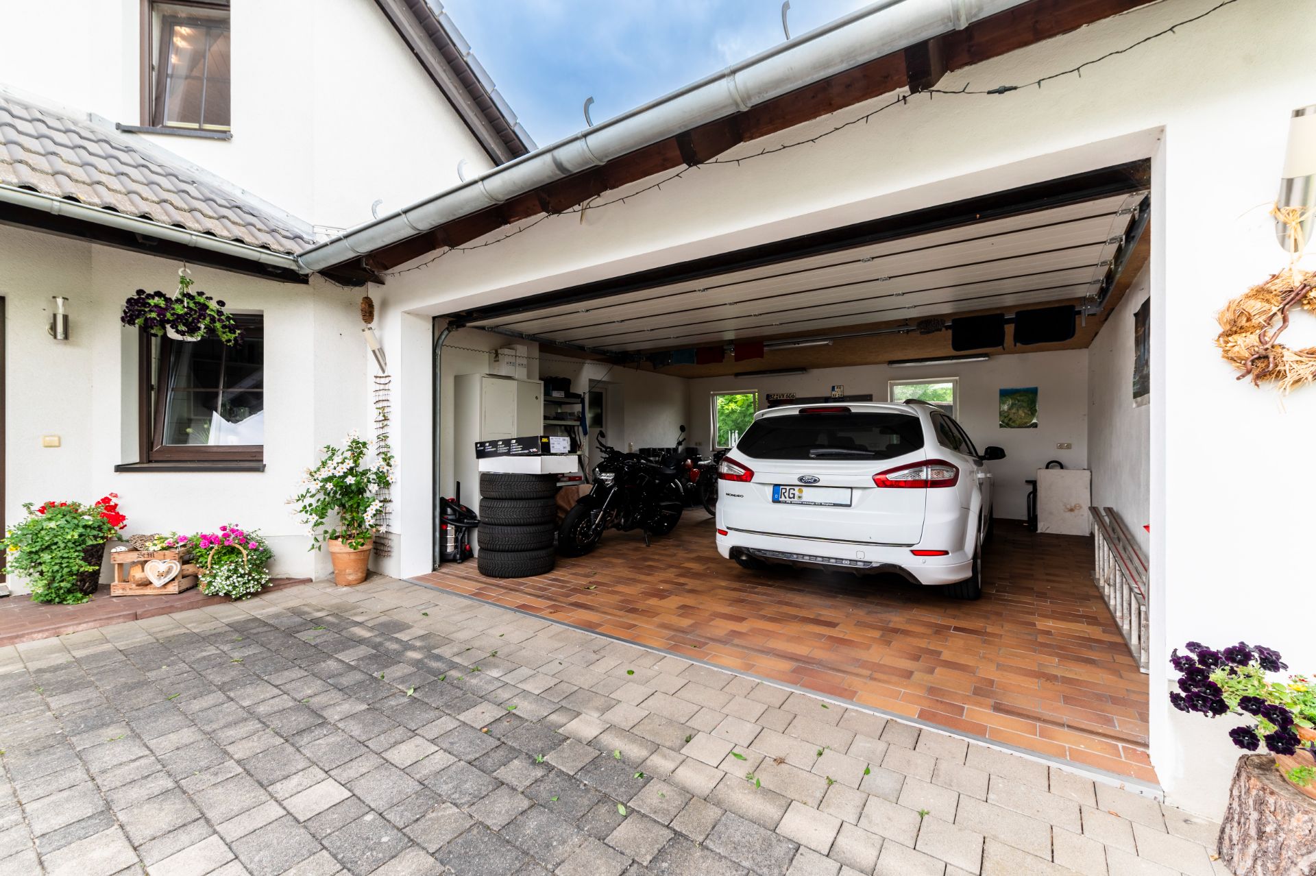 Offene Garage mit weißem Auto, zwei Motorrädern, Regalen und Pflanzen vor einem Wohnhaus.