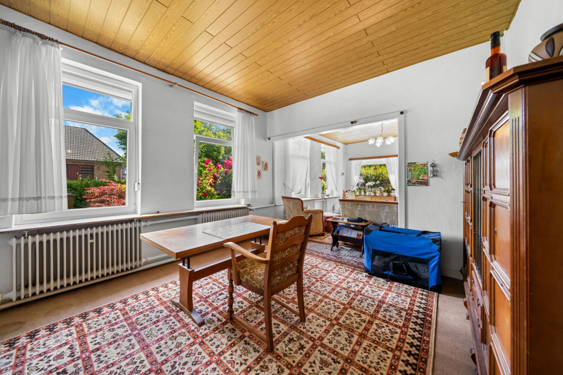Helles Wohnzimmer mit Teppich, Holzmöbeln, großen Fenstern und Blick in einen grünen Garten.