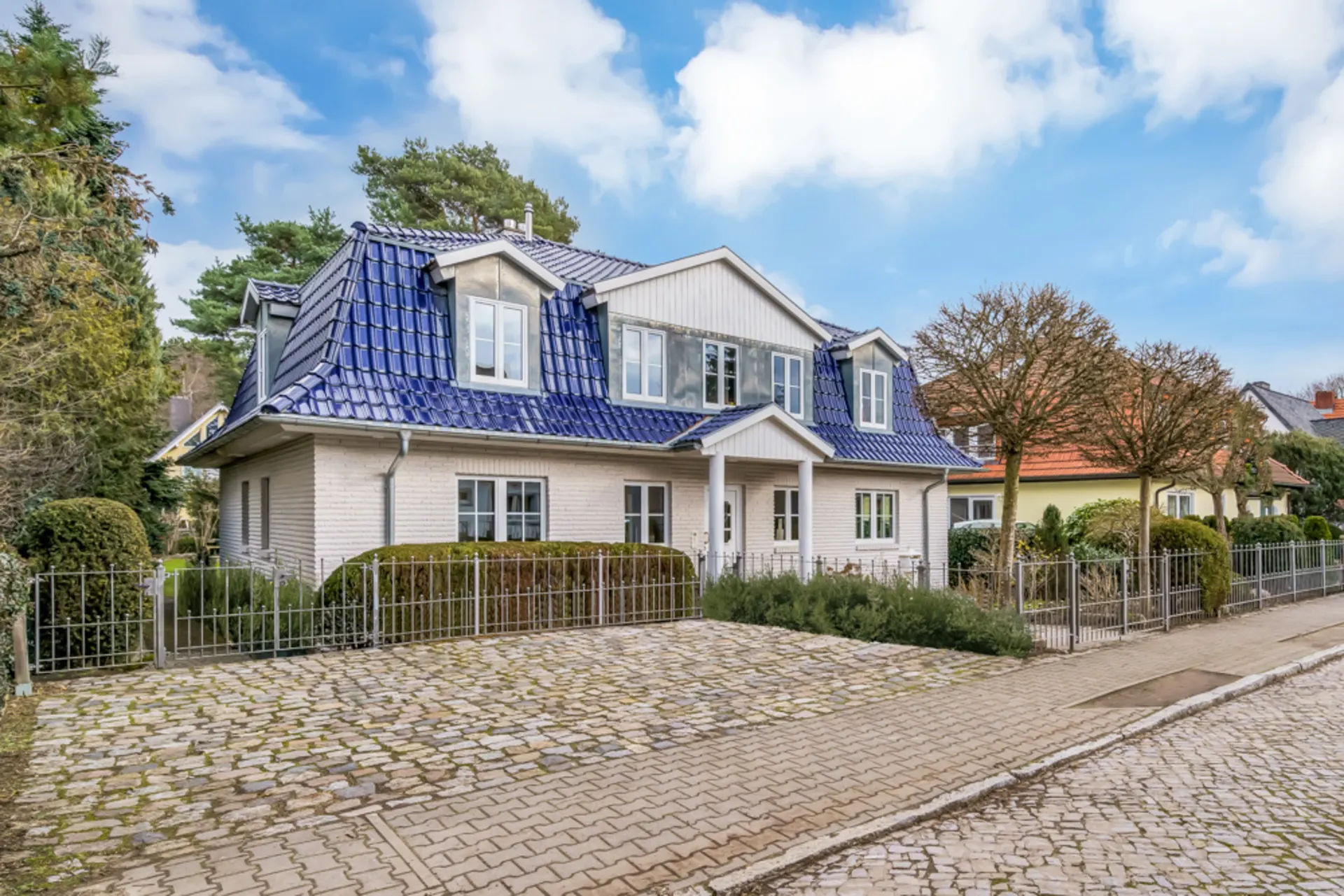 Zweistöckiges Wohnhaus mit blauem Dach, weißer Fassade und kleinem Garten, von Zaun umgeben.