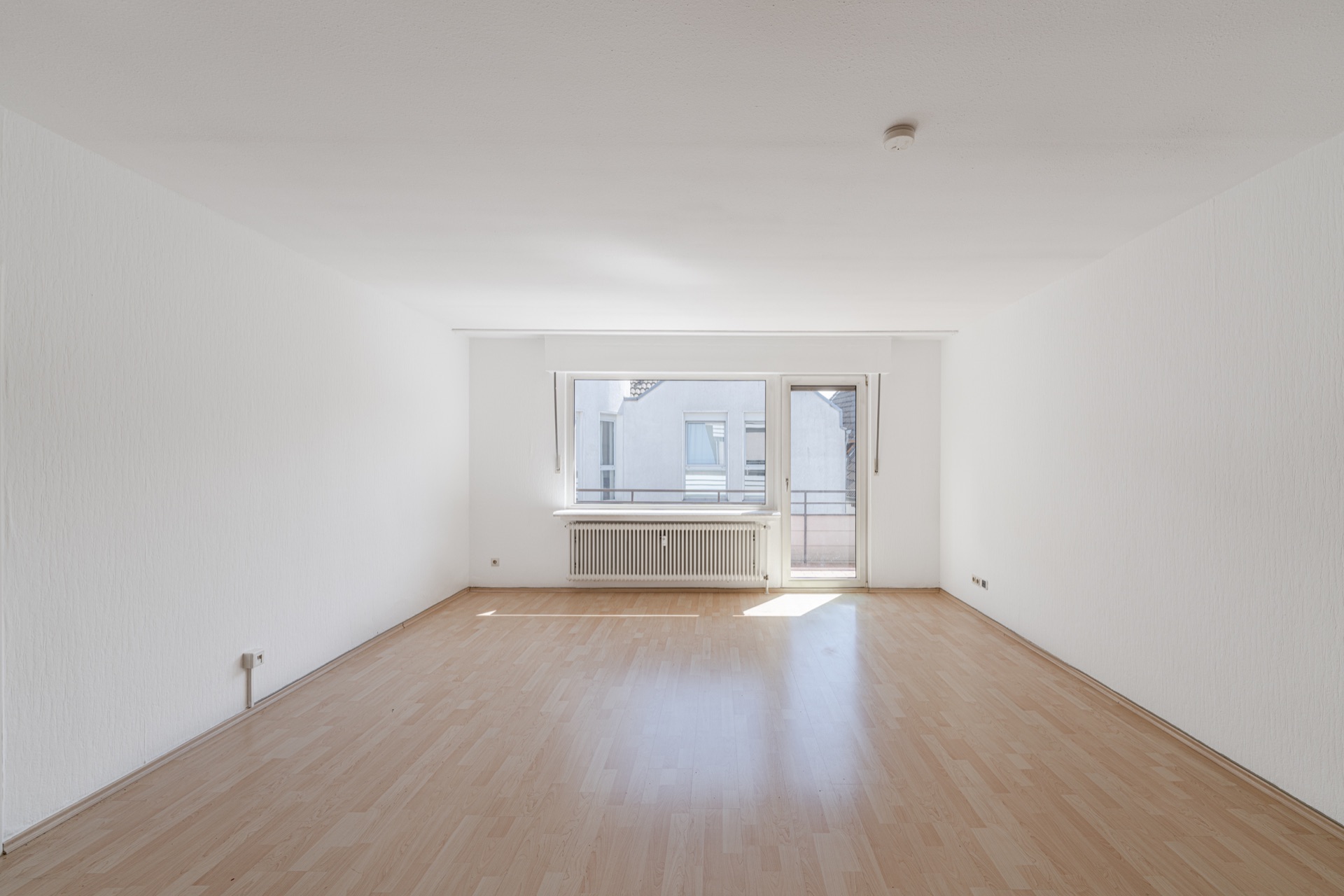 Heller, leerer Raum mit Holzfußboden und großem Fenster samt Tür zum Balkon.
