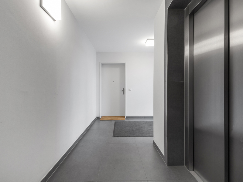 Heller Flur mit Wohnungstür, grauen Fliesen und Aufzug rechts im Bild.