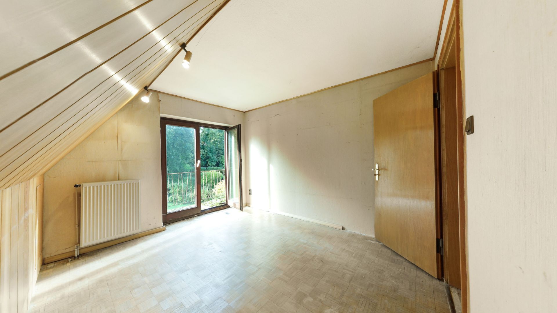 Leeres Zimmer mit schräger Decke, offenem Holztür, Radiator und Glastür zum Balkon.