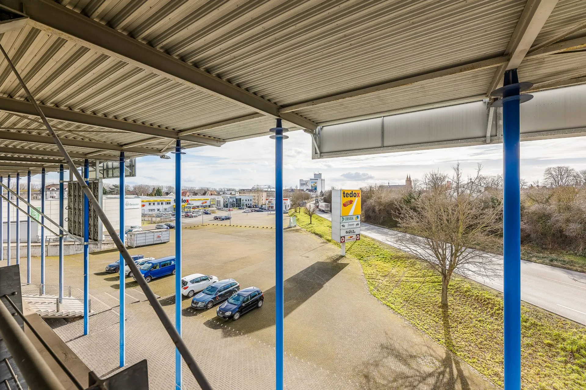 Blick von überdachter Terrasse auf Parkplatz mit mehreren Autos und einigen Bäumen daneben.