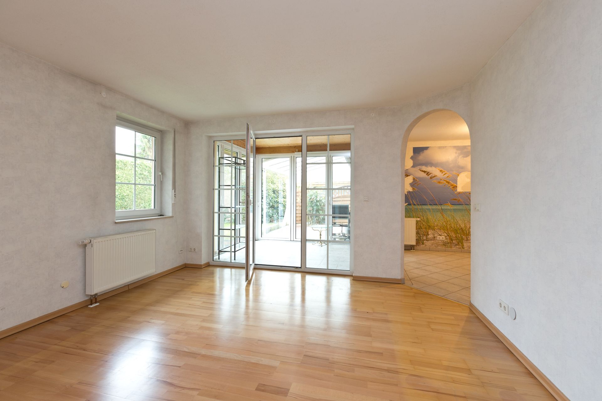 Helles Zimmer mit Parkettboden, Fenster, Glastür zur Terrasse und Durchgang zu weiterem Raum.