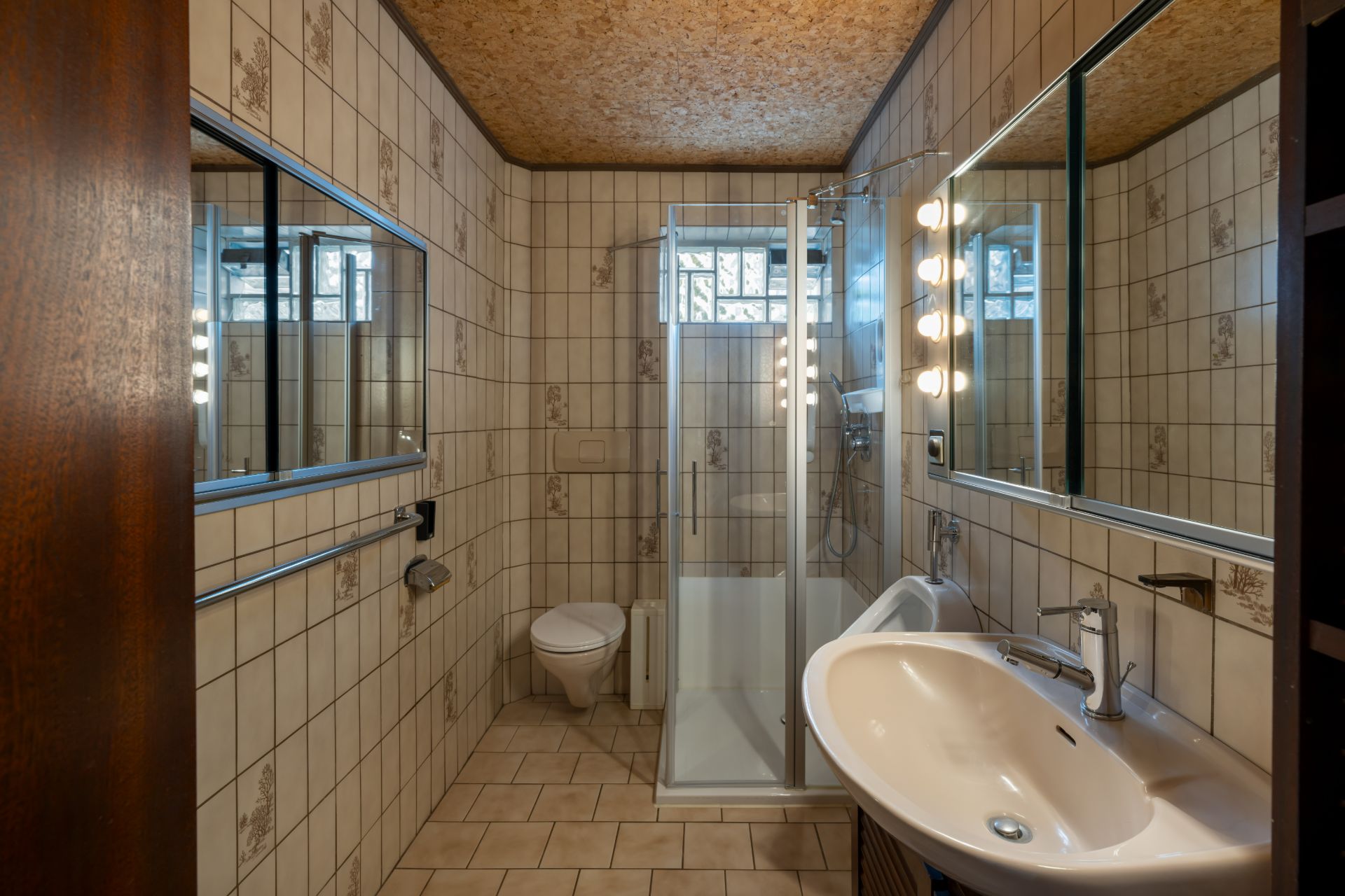 Kleines Bad mit Dusche, WC, Waschbecken und Spiegel, gefliest mit beige-braunen Fliesen.