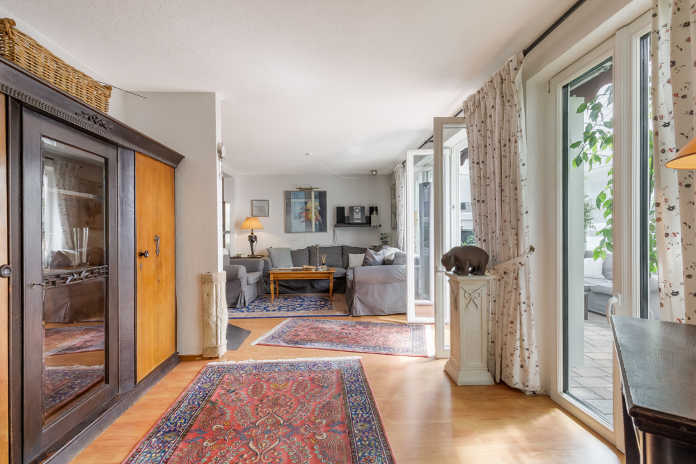 Helles Wohnzimmer mit Teppichen, grauen Sofas, Schrank, vielen Fenstern und Vorhängen.