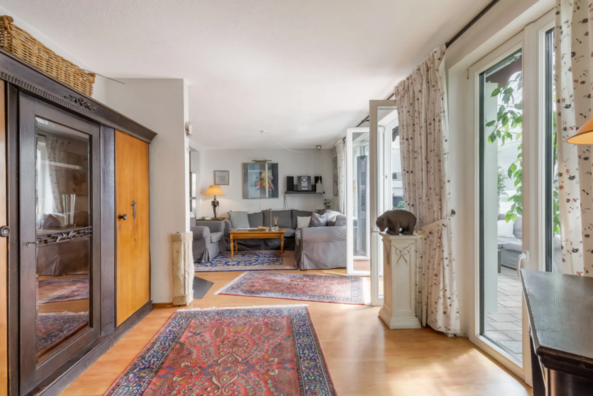 Helles Wohnzimmer mit Teppichen, grauen Sofas, Schrank, vielen Fenstern und Vorhängen.