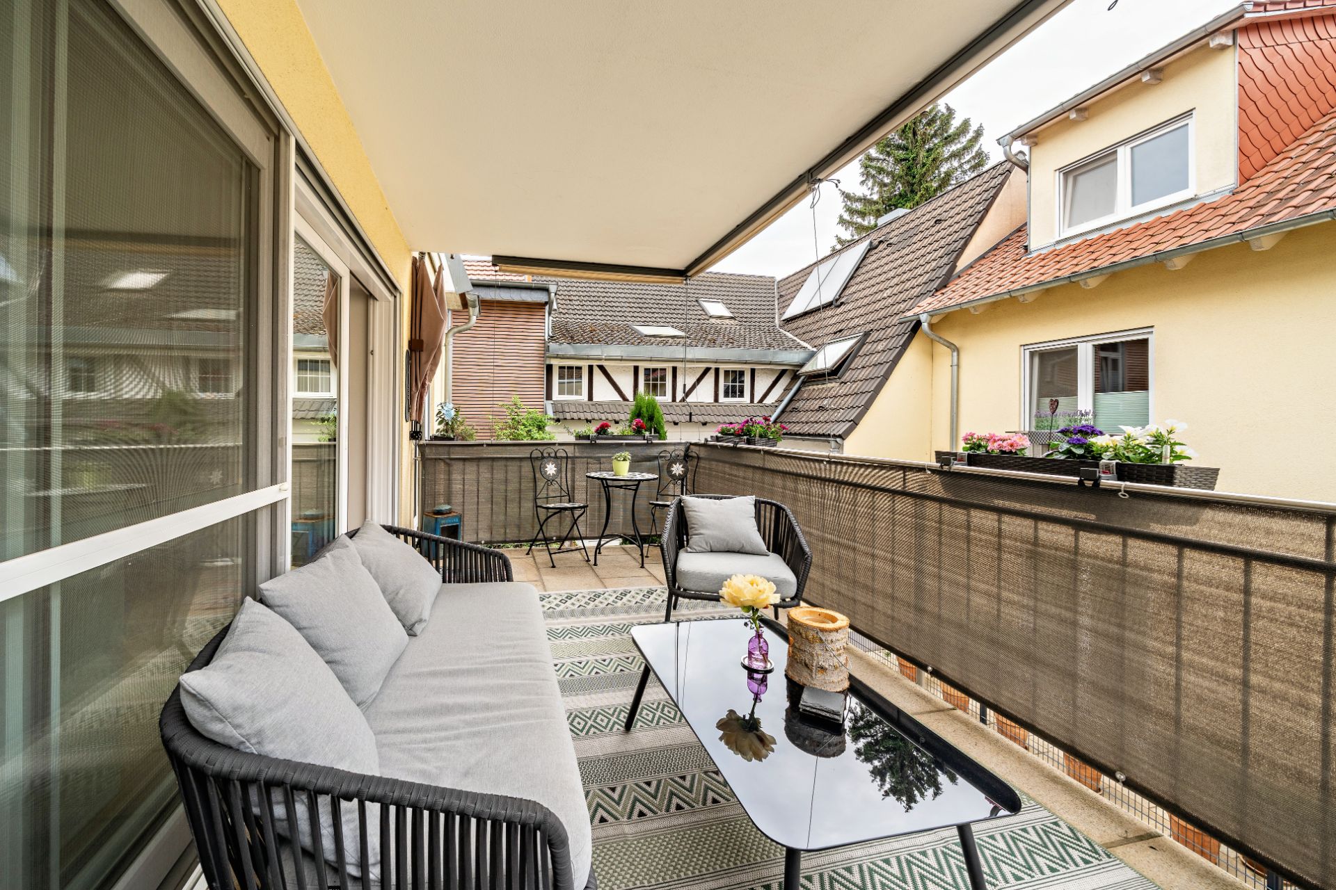Überdachter Balkon mit Sofa, Tisch, Stühlen und Blumenkästen an einem Wohnhaus.
