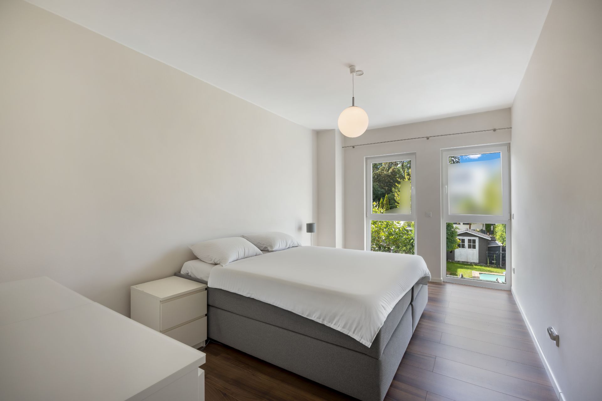Helles Schlafzimmer mit Doppelbett, Nachttisch, Kommode und großem Fenster zum Garten.