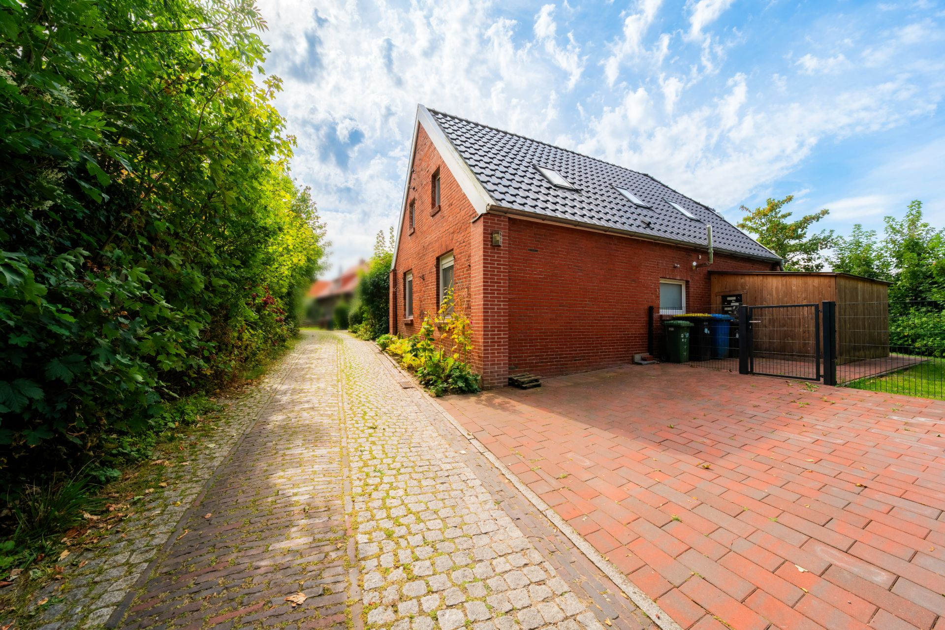 Backsteinwohnhaus mit schrägem Dach, neben gepflastertem Weg und Einfahrt, von Bäumen umgeben.