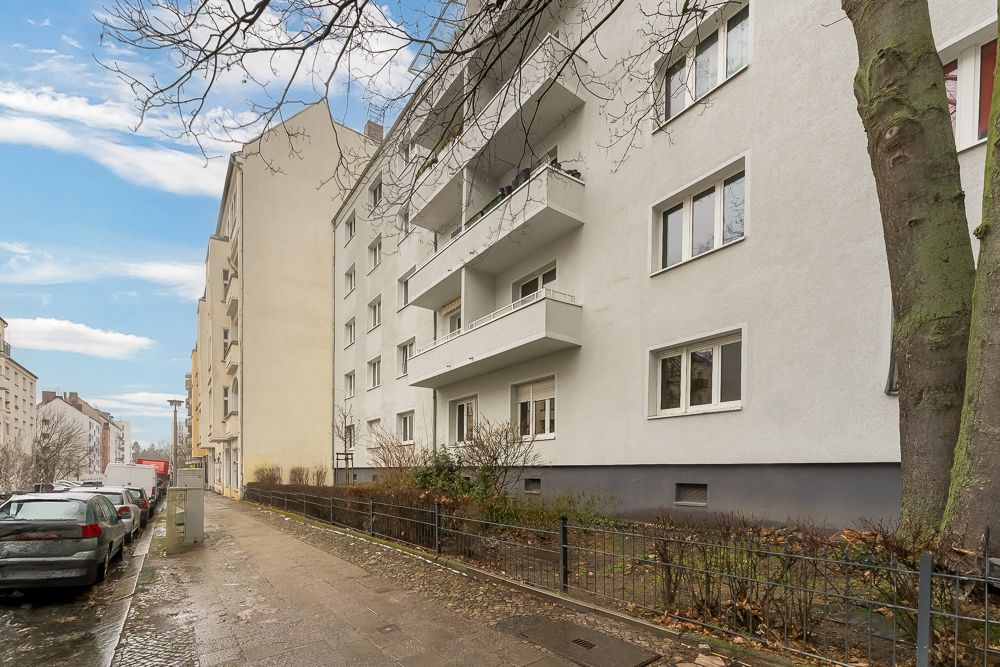 Mehrfamilienhaus mit Balkonen an einer ruhigen Wohnstraße, parkende Autos und Gehweg im Vordergrund.