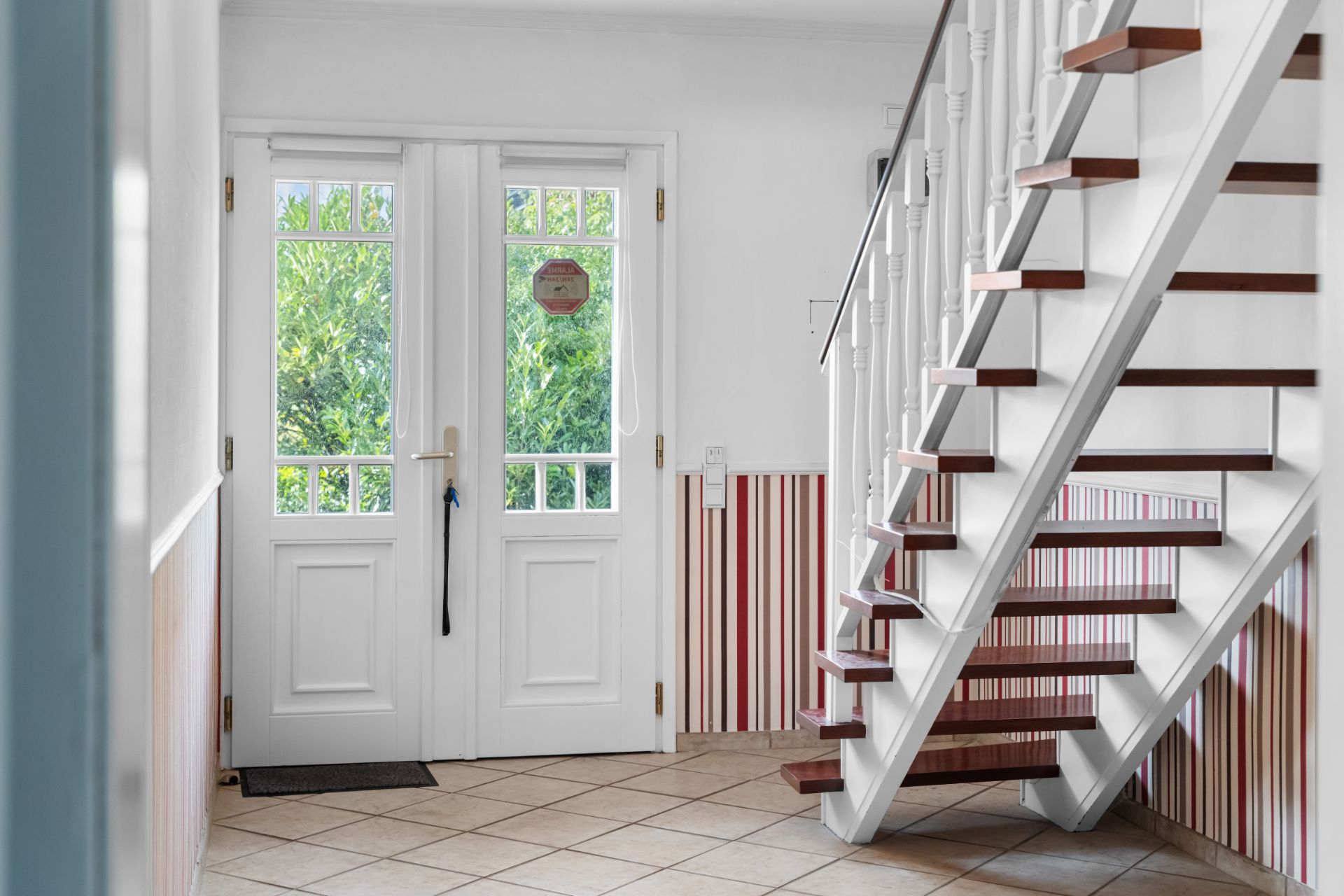 Heller Flur mit weißer Haustür, Fenster und Holztreppe, Fliesenboden und gestreifter Wandverkleidung.