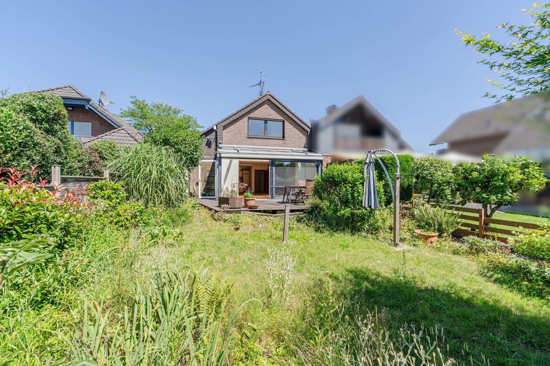 Zweistöckiges Haus mit Terrasse und Garten, umgeben von Pflanzen und Bäumen bei sonnigem Wetter.