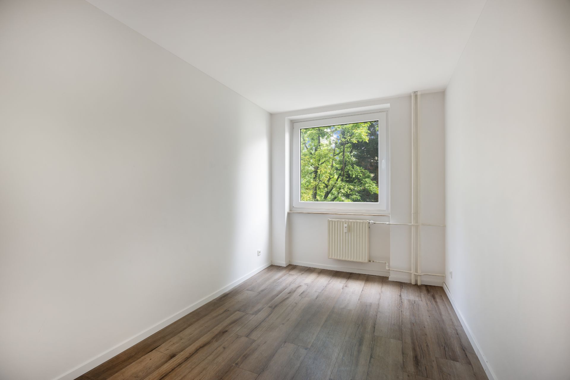 Leerer, heller Raum mit Holzboden, weißem Fenster und Blick ins Grüne.