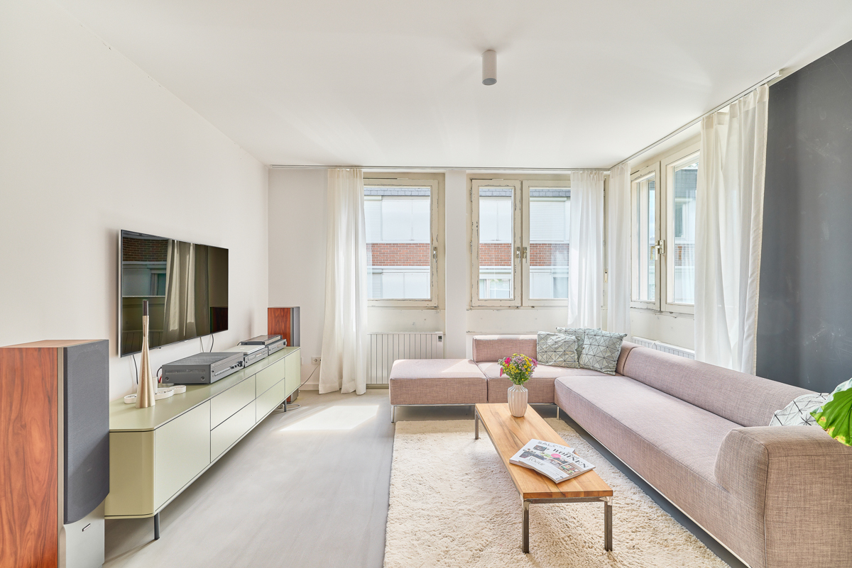 Helles Wohnzimmer mit Ecksofa, Couchtisch, TV und großen Fenstern mit weißen Vorhängen.