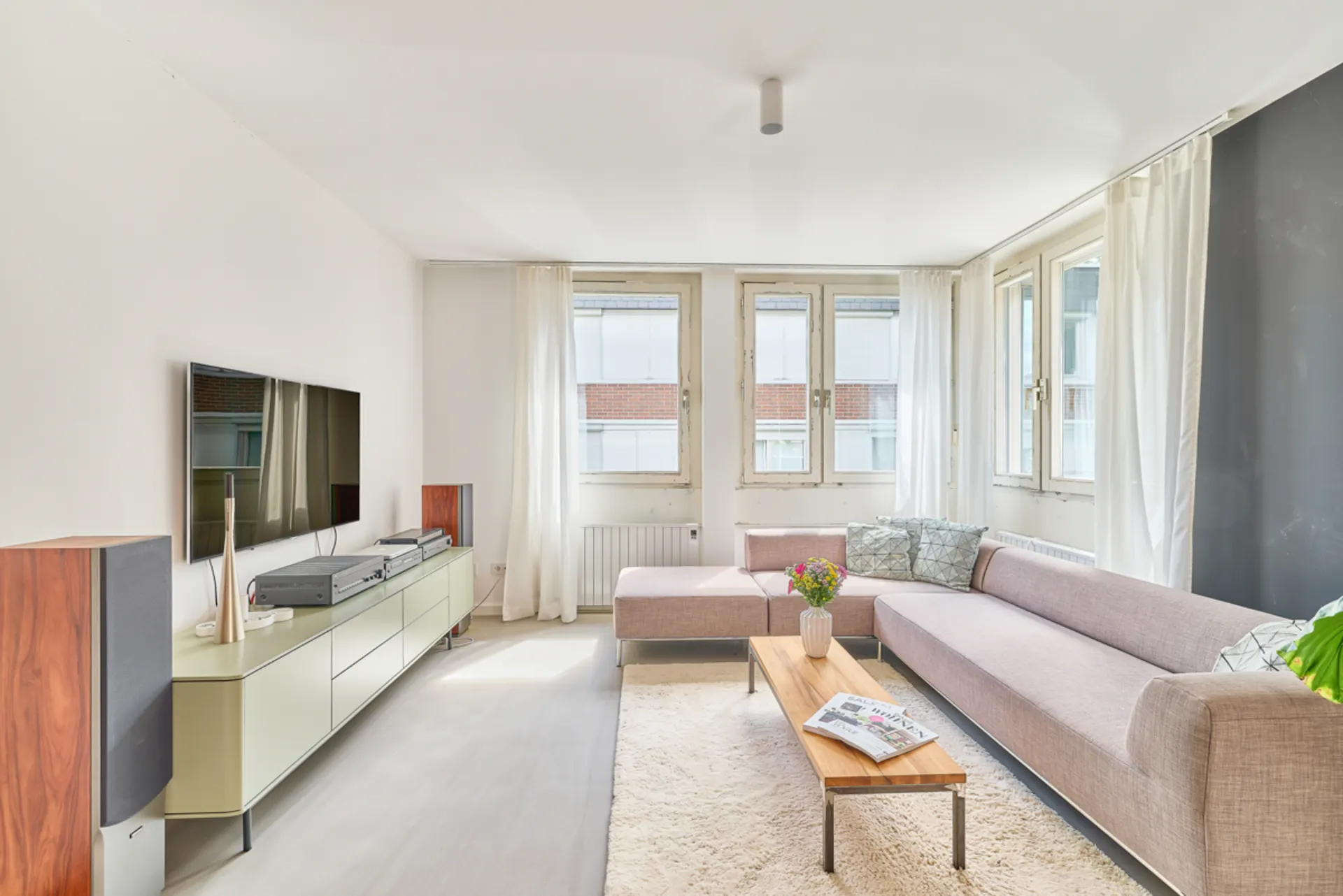 Helles Wohnzimmer mit Ecksofa, Couchtisch, TV und großen Fenstern mit weißen Vorhängen.