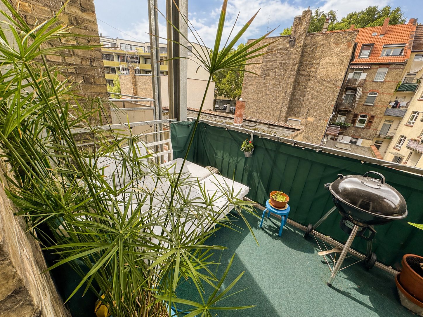 Kleiner Balkon mit Pflanzen, einem Grill, Tisch und Blick auf benachbarte Wohngebäude.