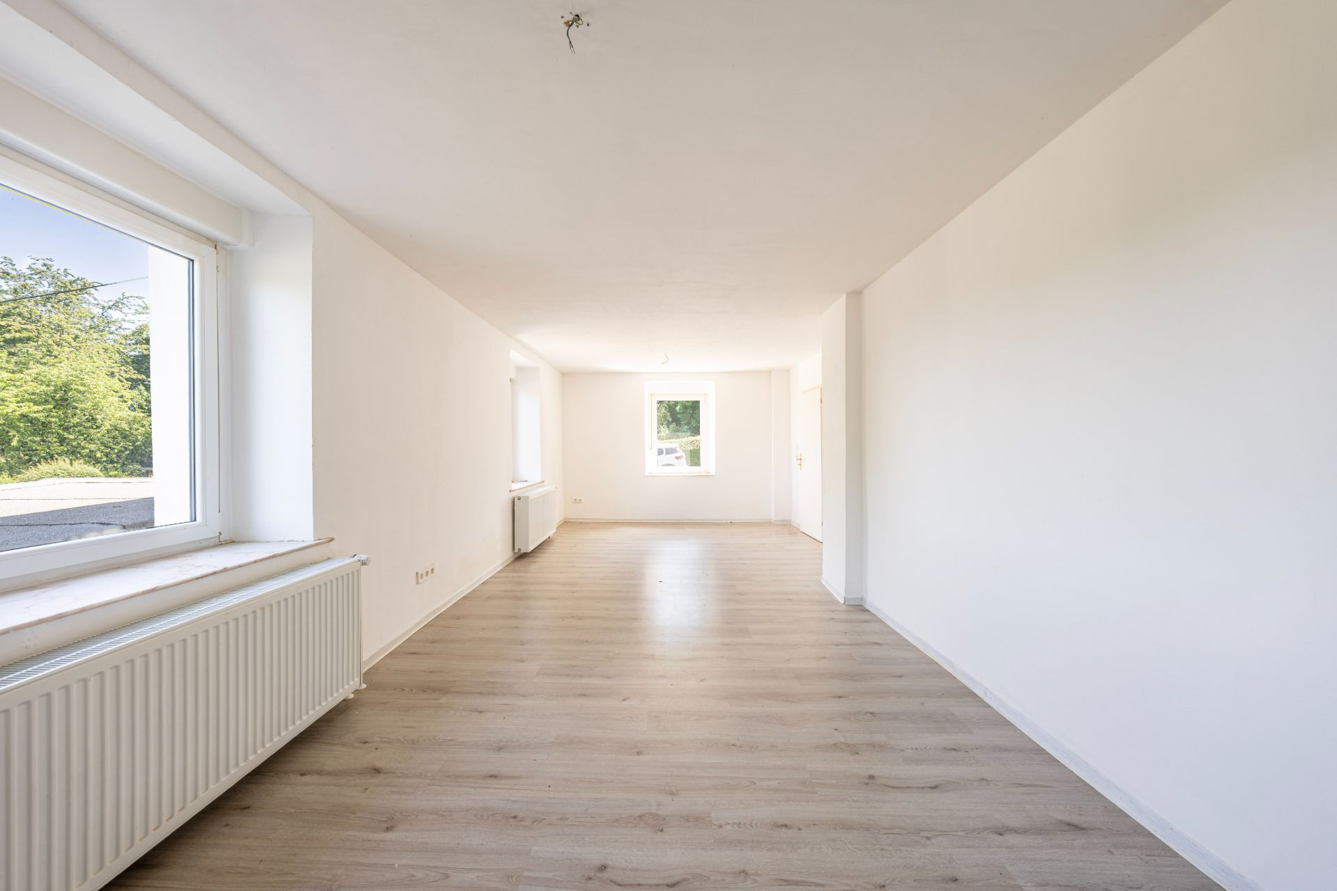 Helles, leeres Zimmer mit zwei großen Fenstern, weiß gestrichenen Wänden und hellen Holzböden.