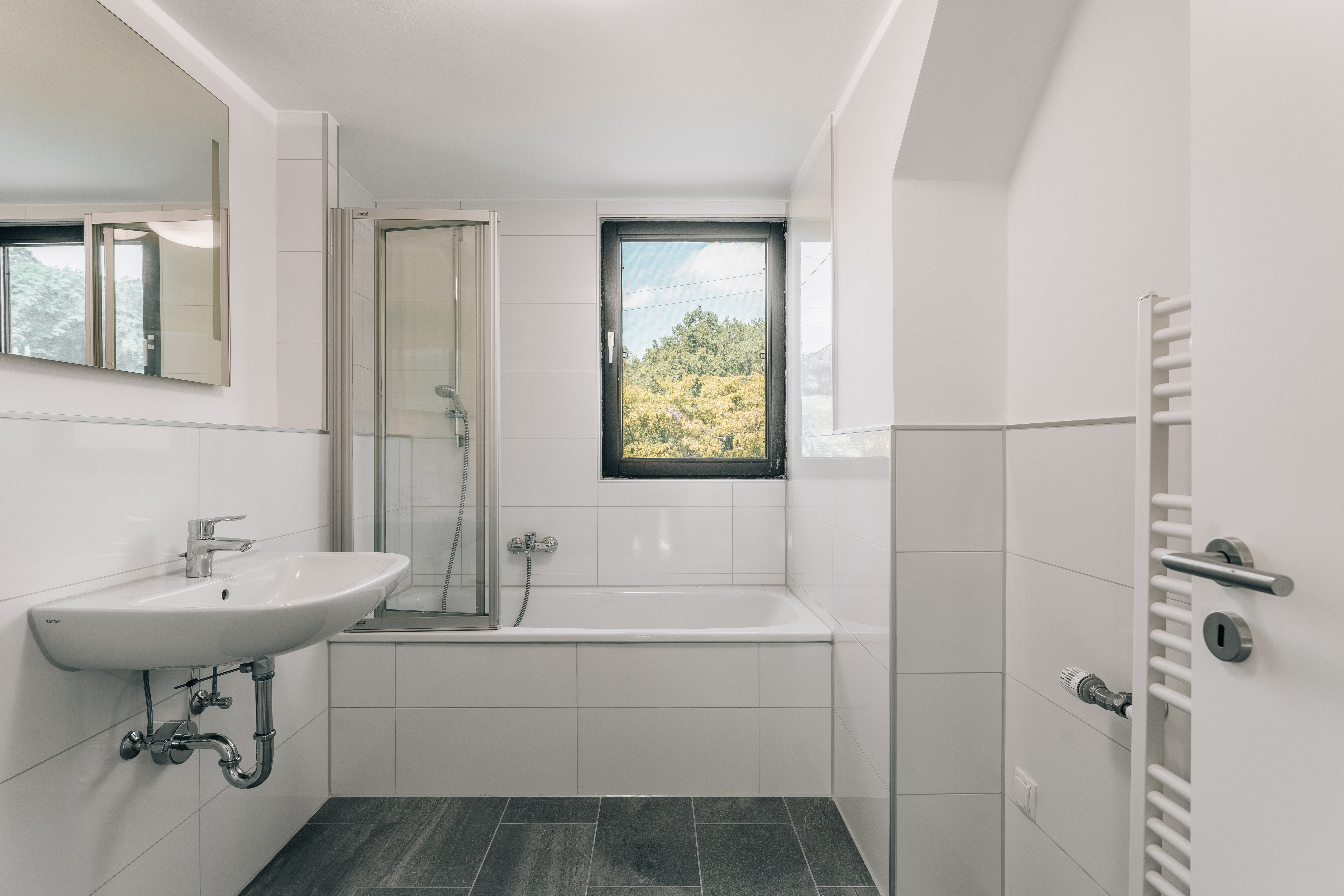 Helles Badezimmer mit Fenster, Badewanne, Waschbecken und Handtuchheizkörper.