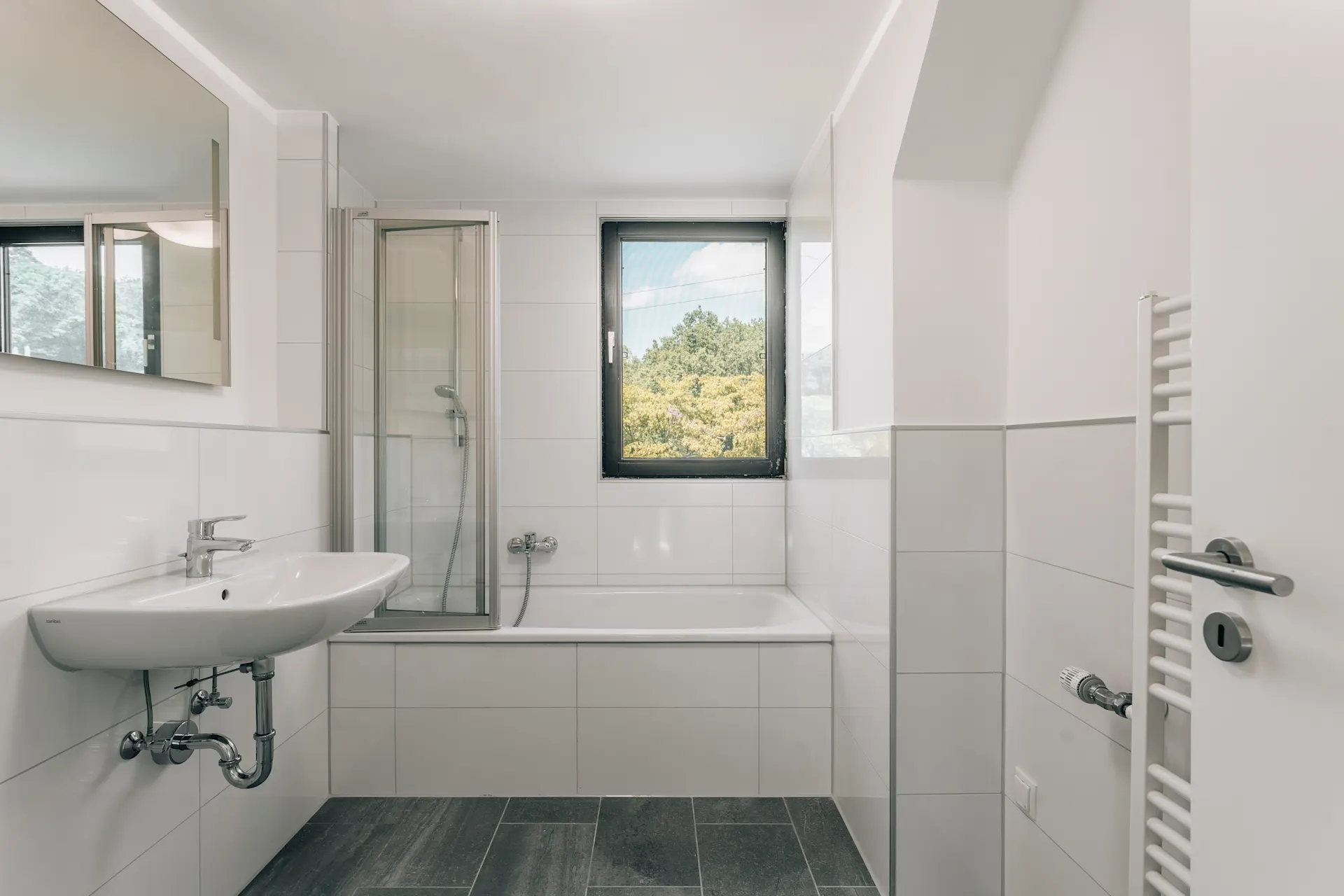 Helles Badezimmer mit Fenster, Badewanne, Waschbecken und Handtuchheizkörper.