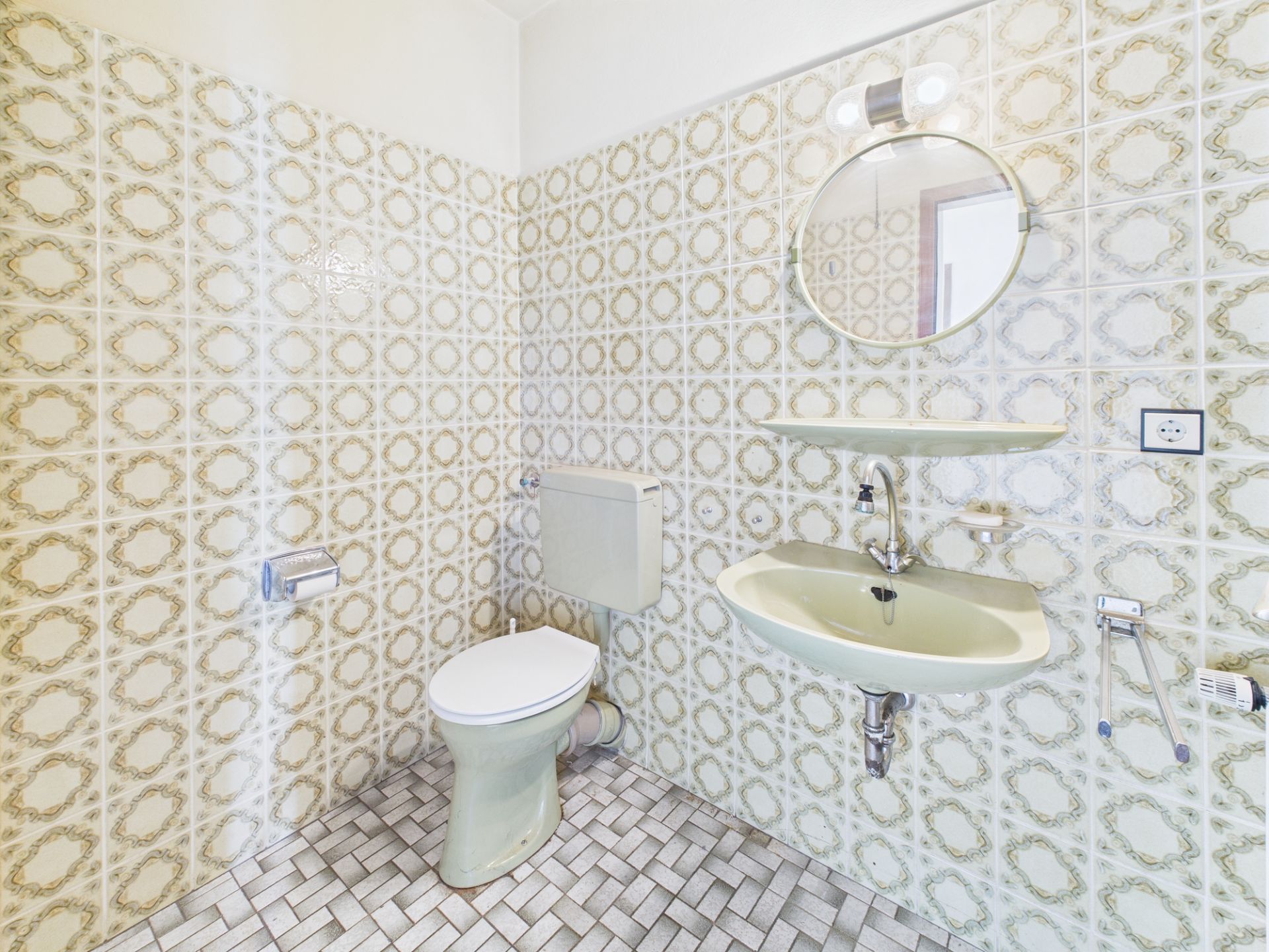 Kleines Badezimmer mit Toilette, Waschbecken und rundem Spiegel, Fliesen mit Muster.