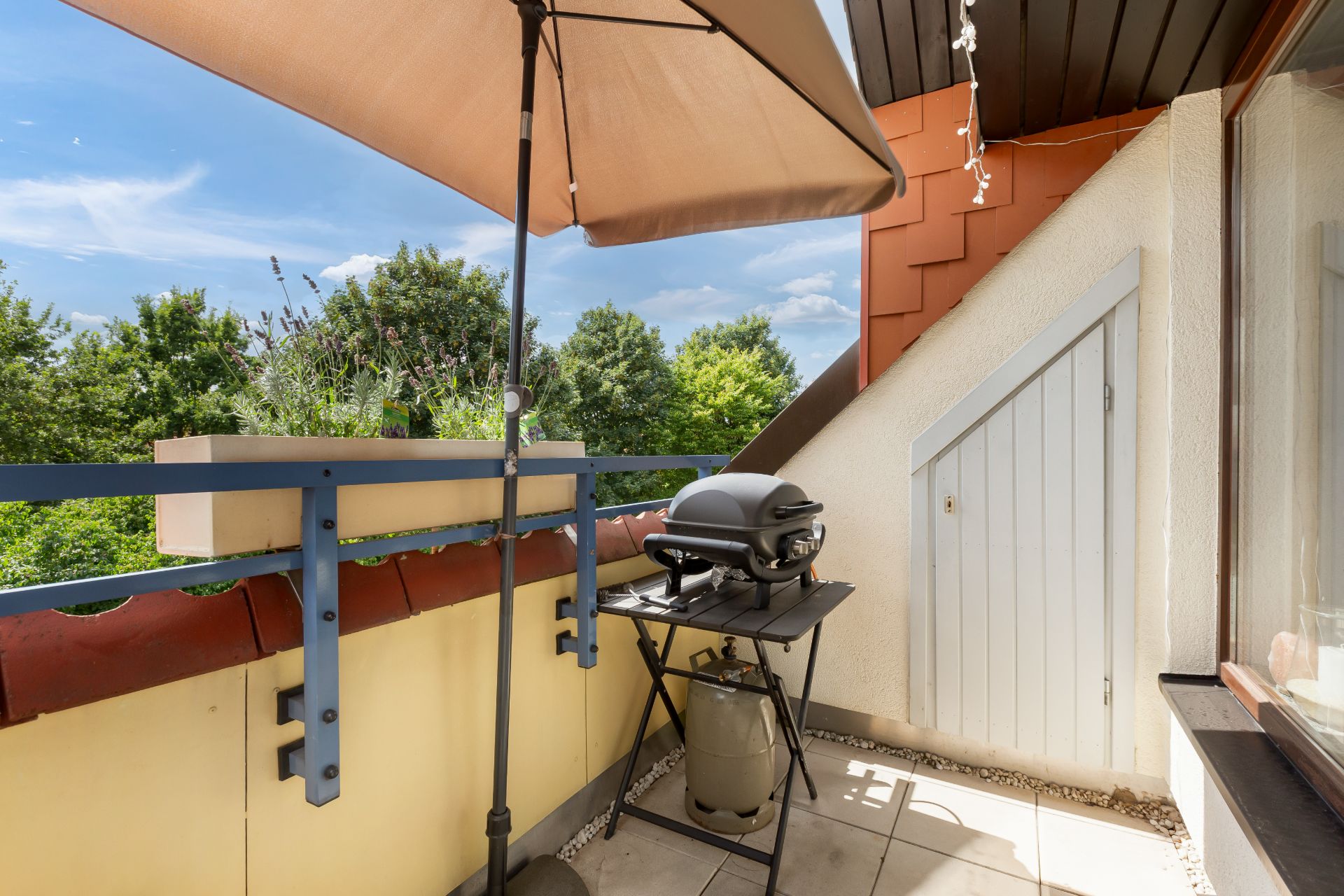 Kleiner Balkon mit Grill, Sonnenschirm, Pflanzkasten und seitlicher Tür, Ausblick auf Bäume.