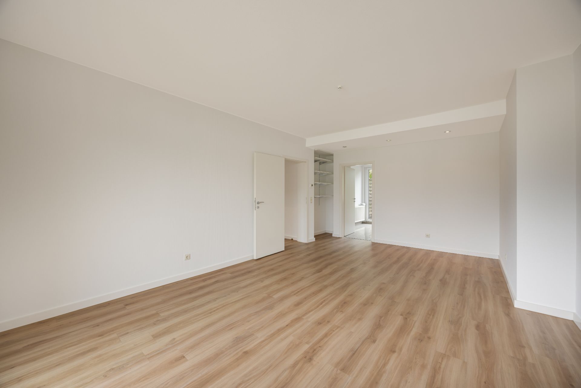 Helles, leeres Zimmer mit Holzfußboden, weißen Wänden und einer offenen Tür.