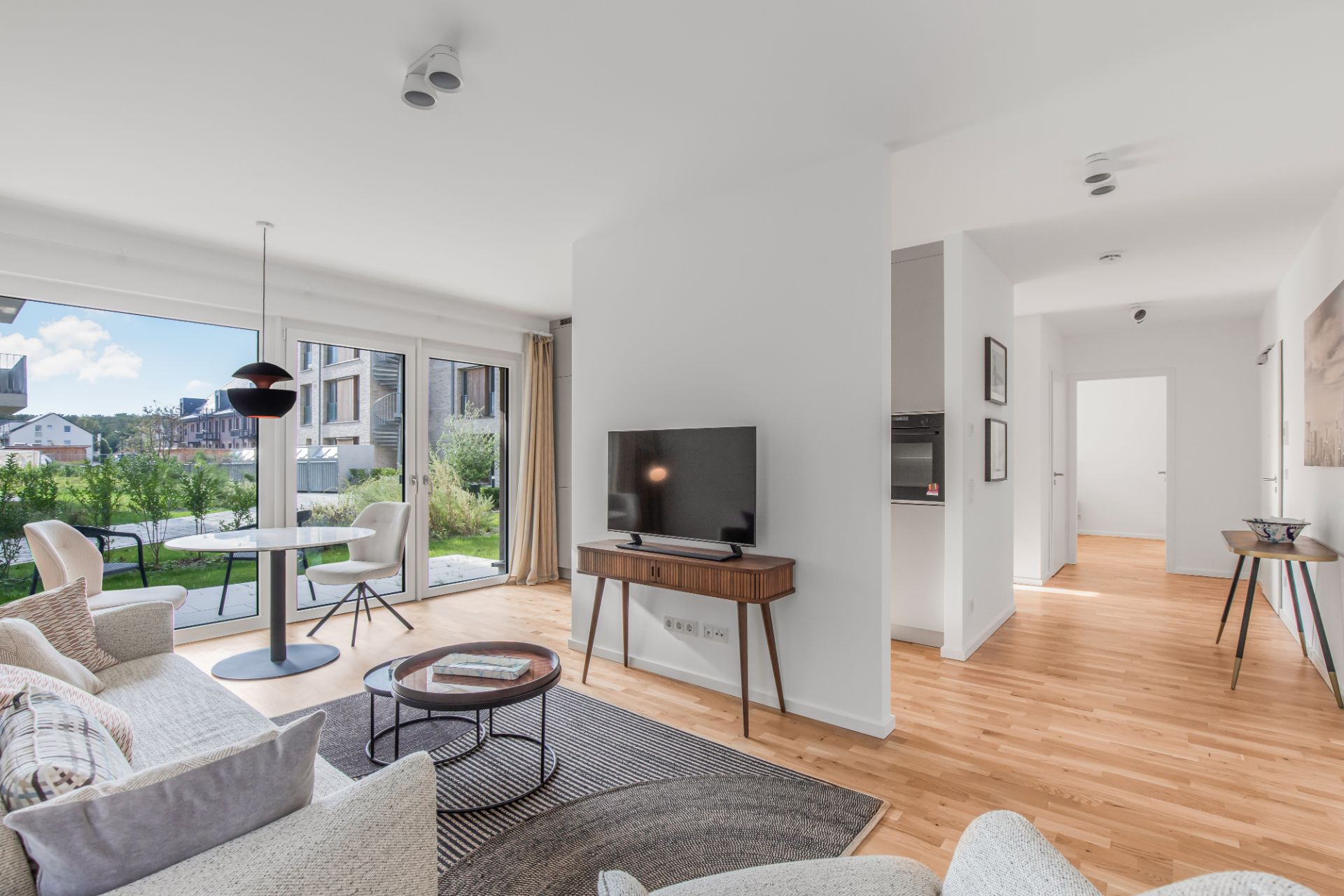 Helles Wohnzimmer mit Sofa, Fernseher, Esstisch und Blick auf einen Garten durch große Fenster.