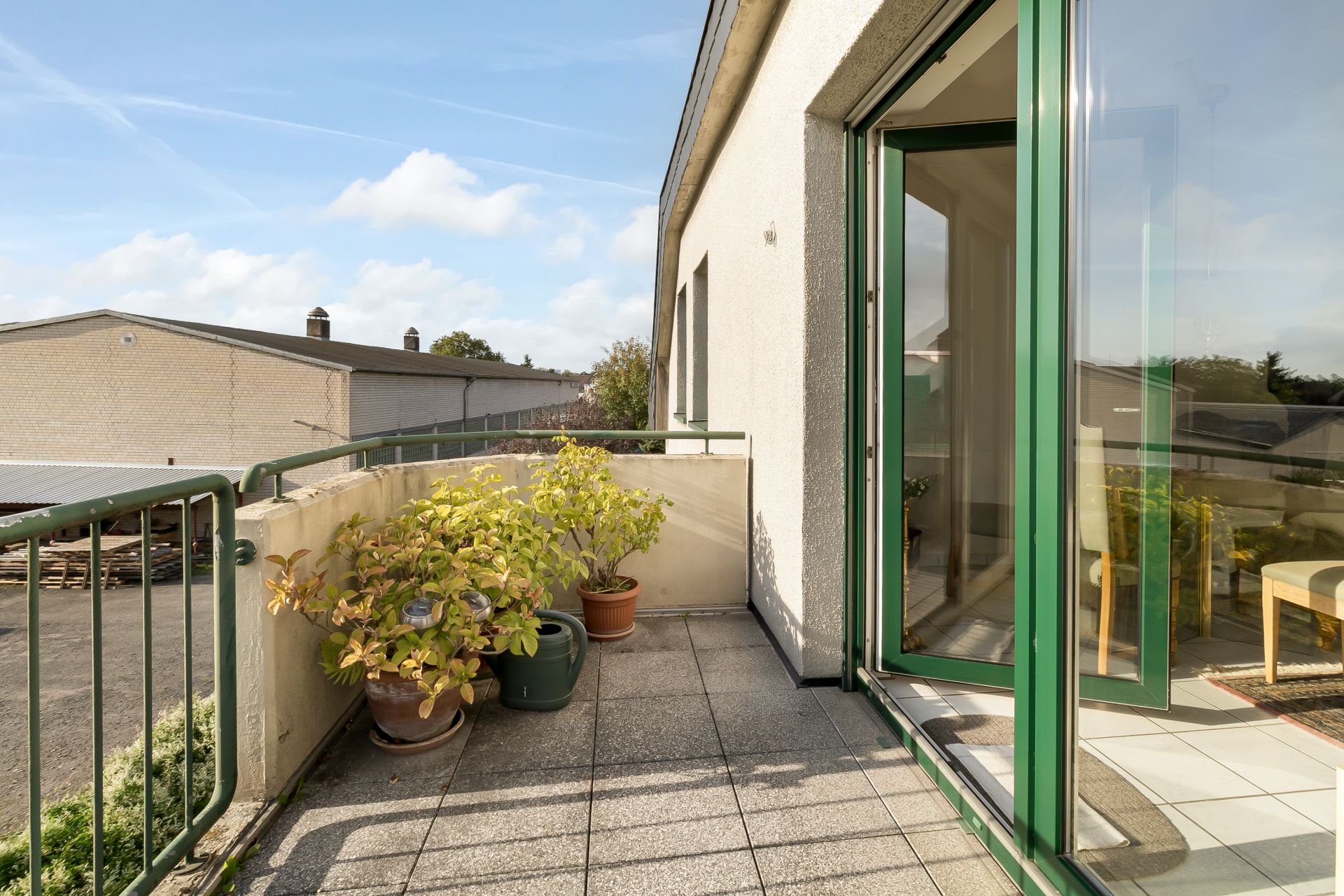 Balkon mit mehreren Pflanzen in Töpfen und offener grüner Tür zu einem hellen Raum.