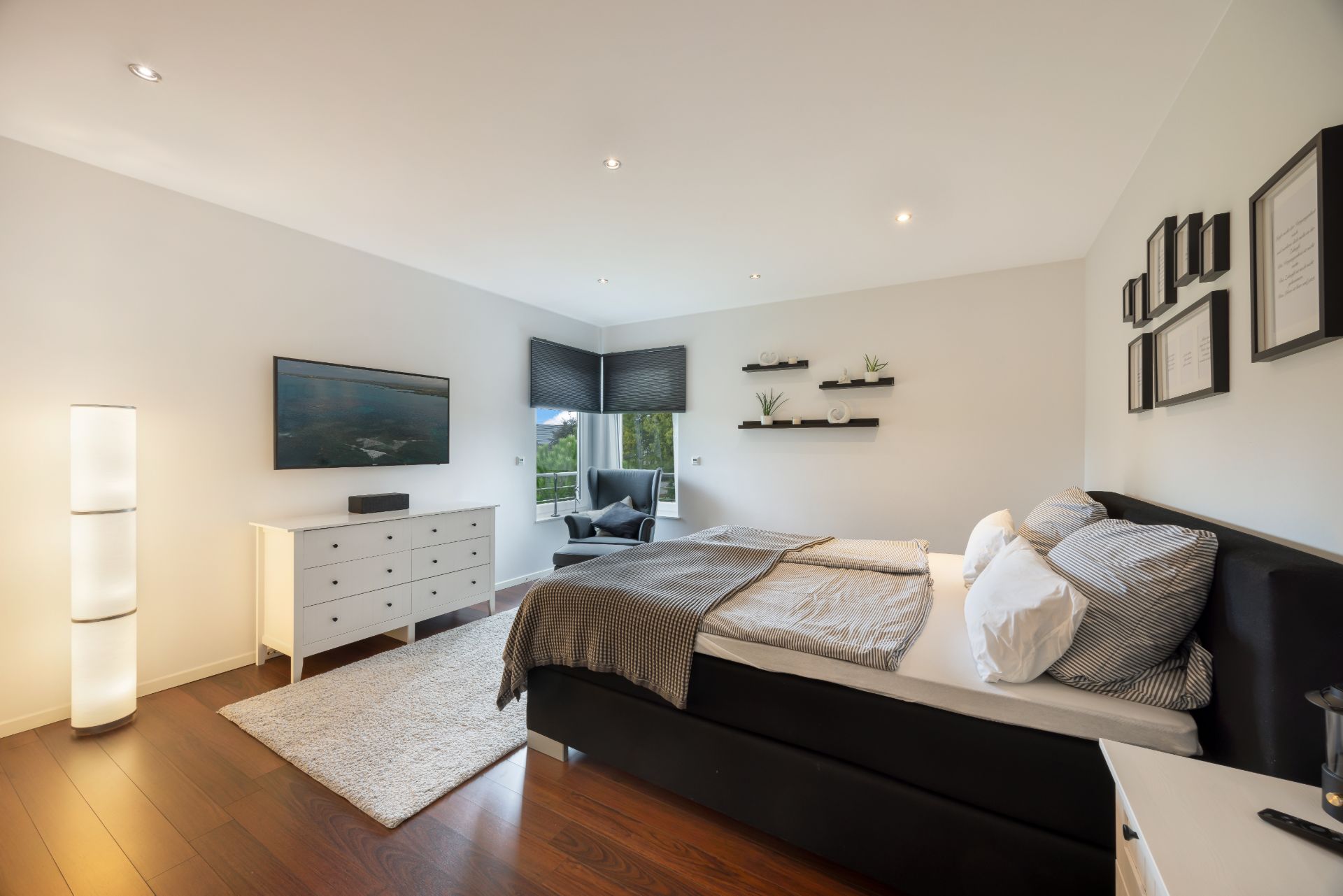 Modernes Schlafzimmer mit Doppelbett, Sessel am Fenster, Kommode, Teppich und Wandregalen.