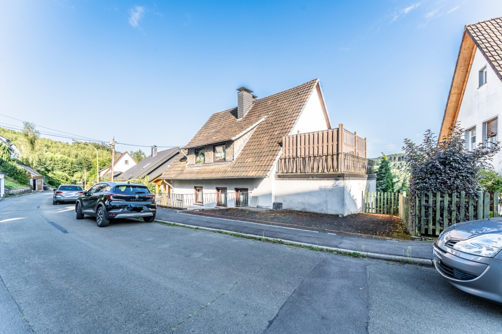 Einfamilienhaus mit schrägem Dach und Holzzaun an einer ruhigen Straße mit geparkten Autos.