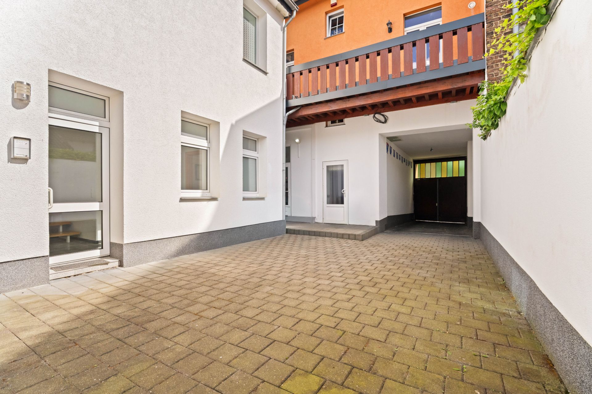 Gepflasterter Innenhof mit weißen Wänden, Fenstern und einer Tür, überdacht durch einen Balkon.