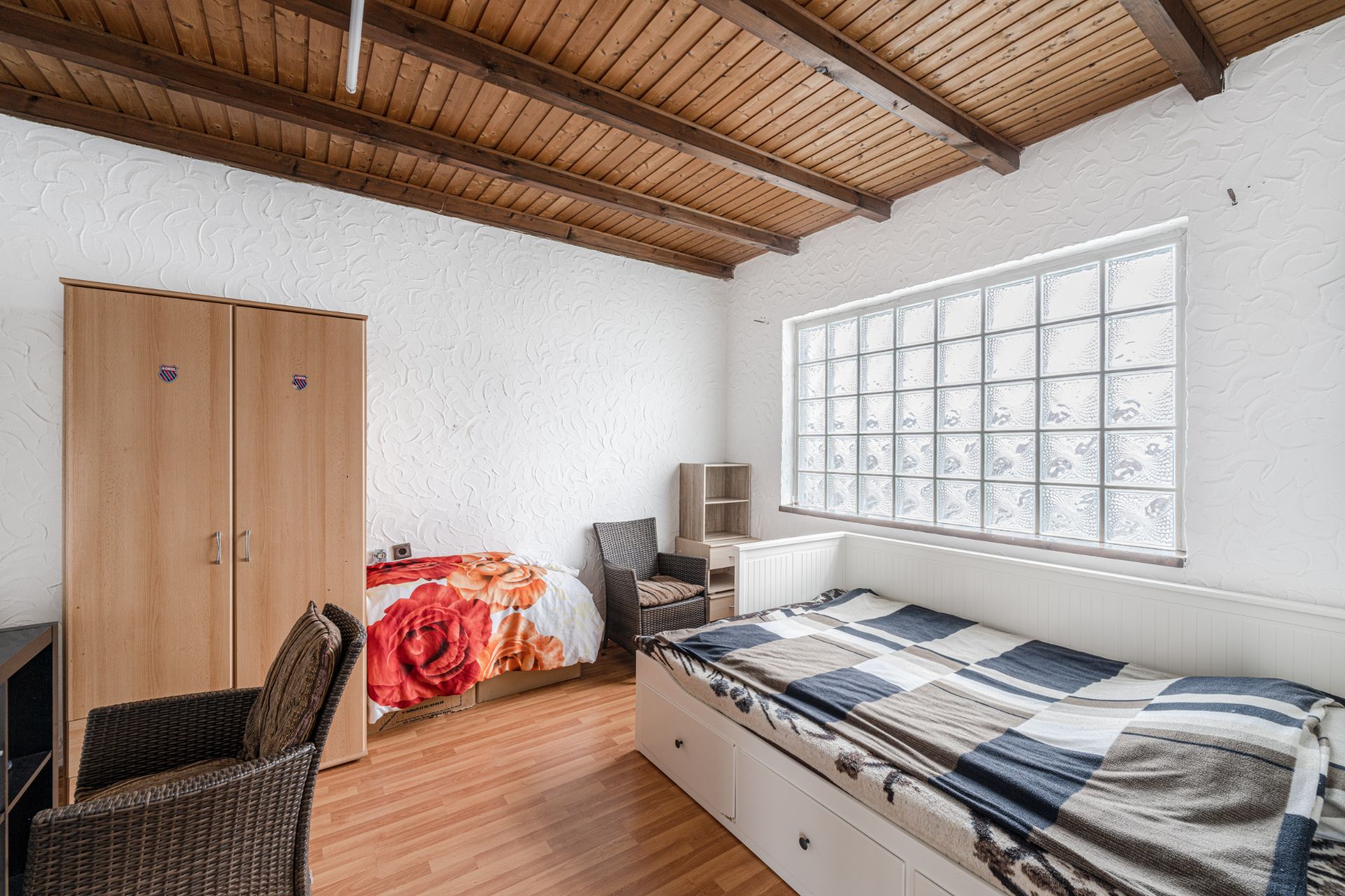 Helles Schlafzimmer mit zwei Betten, Holzdecke, Schrank und großem Fenster aus Glasbausteinen.