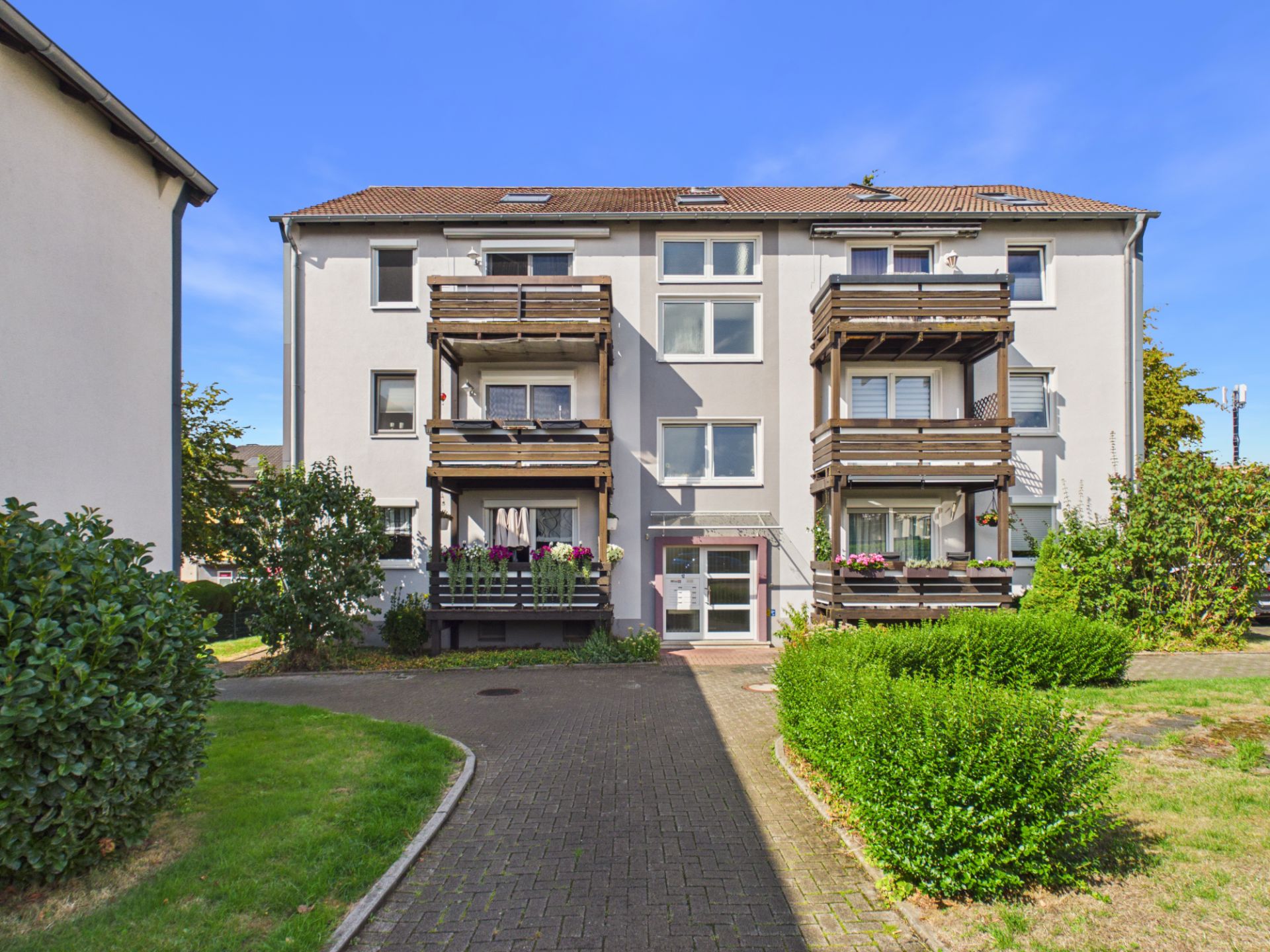 Drei­stöckiges Wohnhaus mit zwei Holzbalkonen und Vorgarten, gepflasterter Weg zum Eingang.